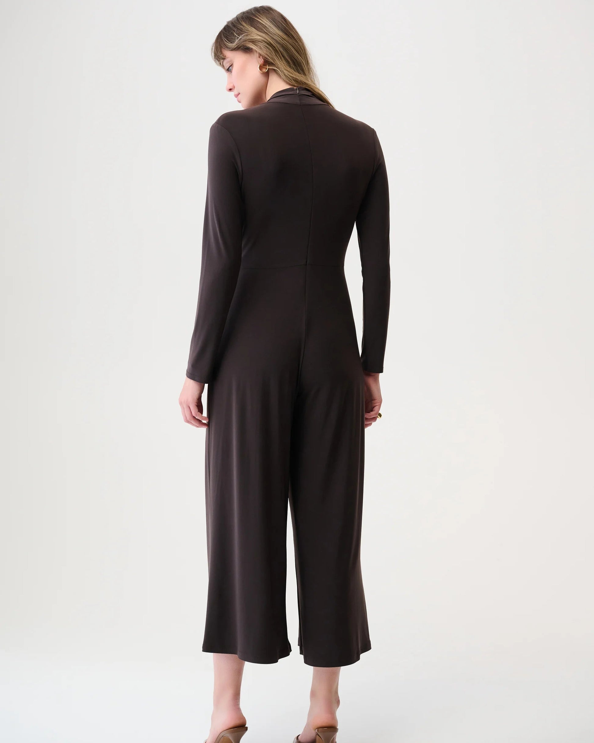 Back view of a woman wearing a dark brown long-sleeve jumpsuit on a white background