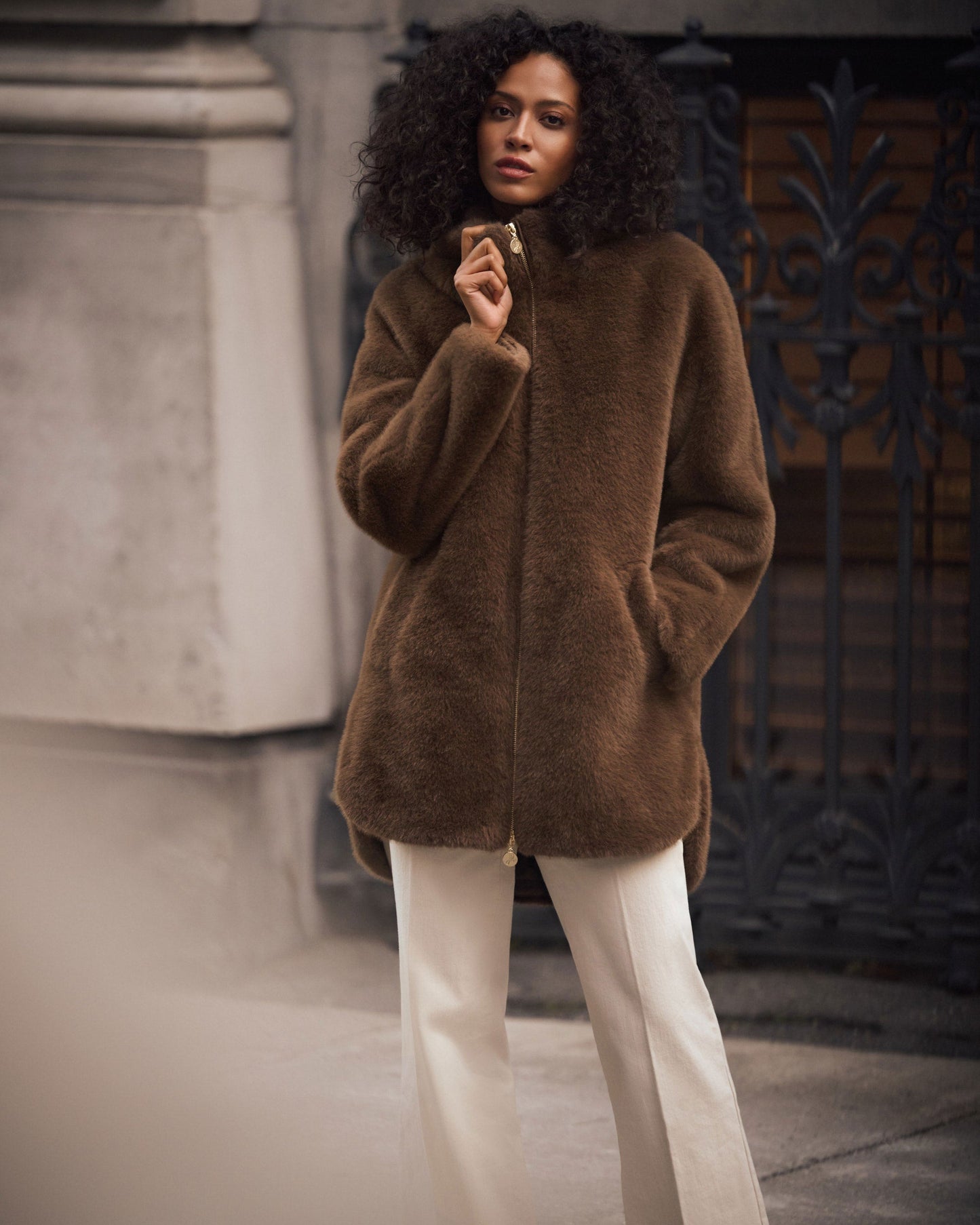 Woman wearing a brown coat and white pants standing in an urban setting.