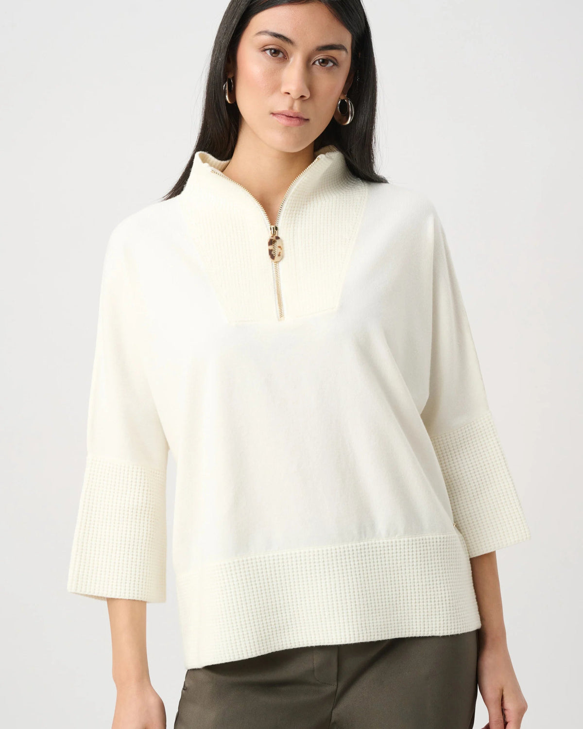 A woman modeling a cream-colored textured knit top with a half-zip neckline, three-quarter sleeves, and a ribbed hem, paired with olive green pants against a light gray studio background.