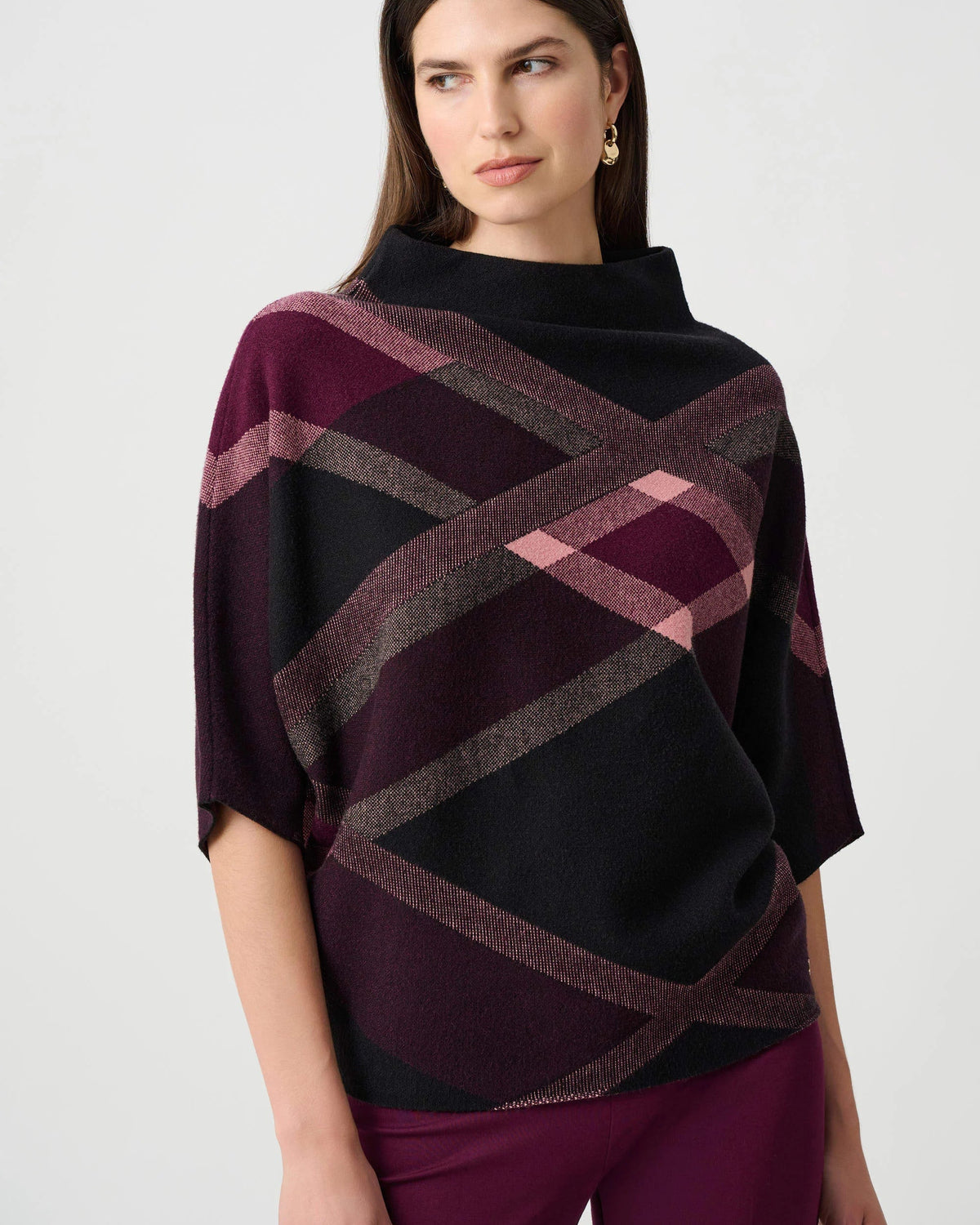 A woman modeling a short-sleeve knit sweater with a geometric plaid pattern in black, deep burgundy, and dusty pink, paired with burgundy pants against a light gray studio background.
