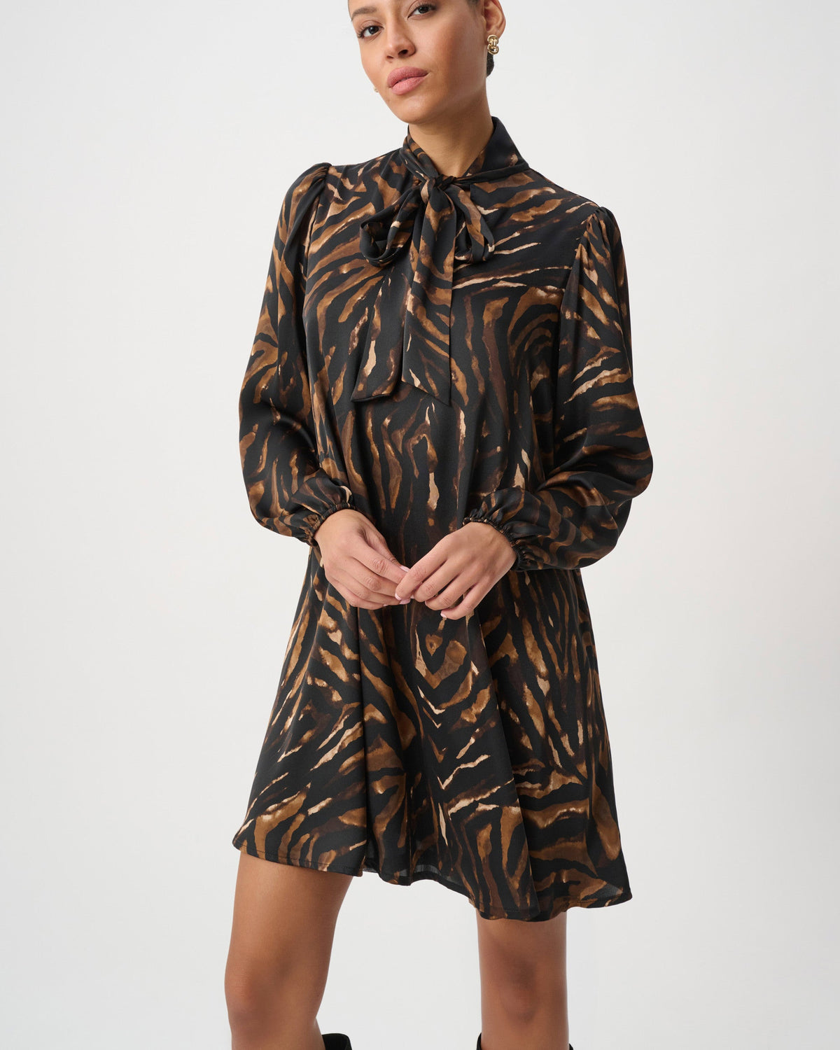 A woman modeling a long-sleeve black dress with a brown and gold zebra-like abstract print, featuring a tie-neck detail and a flared skirt, paired with black knee-high boots against a light gray studio background.