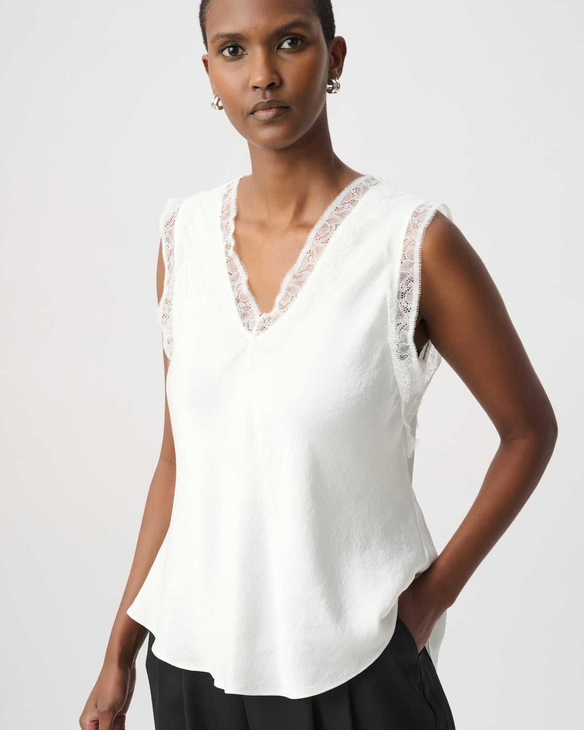 A woman modeling a sleeveless white top with a lace V-neck trim, paired with black pants, against a light gray studio background.