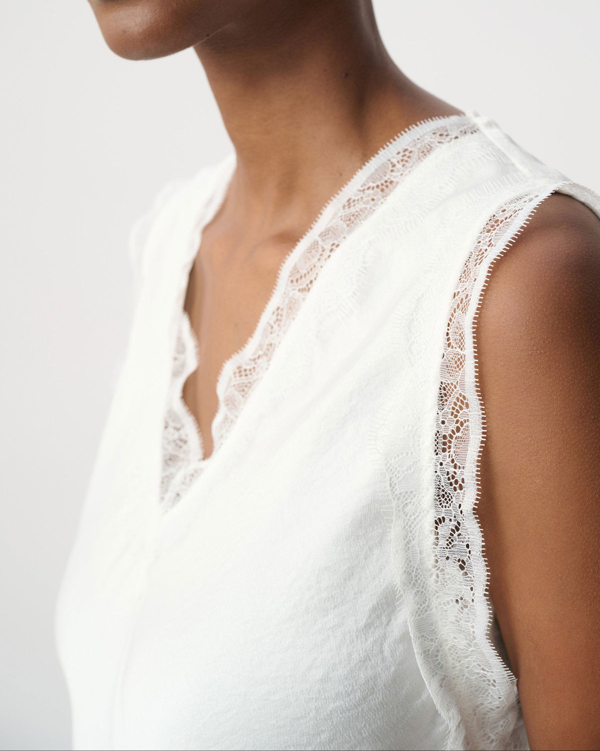 Close-up of a model wearing a white sleeveless top with delicate lace trim along the neckline and armholes, paired with silver hoop earrings against a light gray background.
