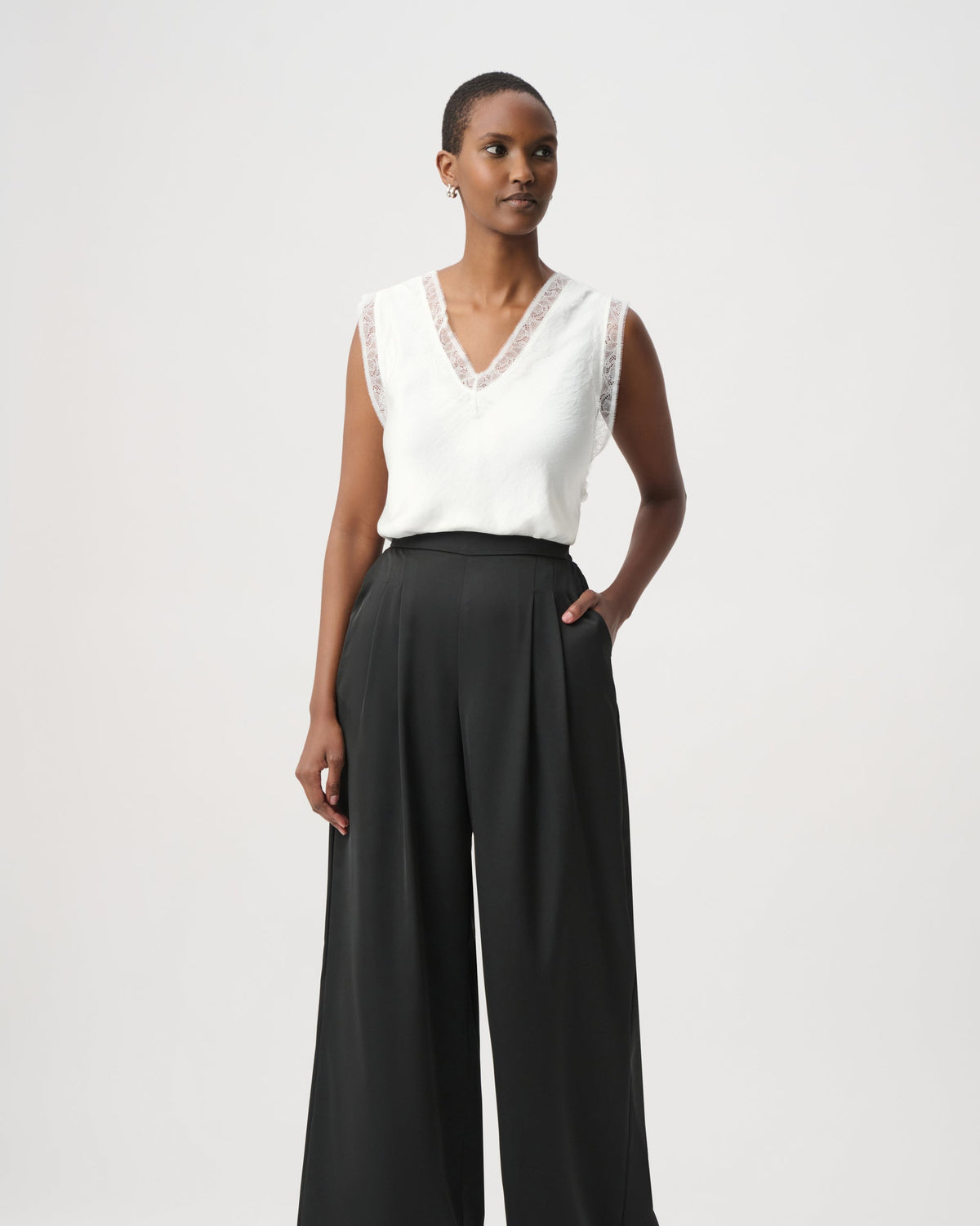 A model wearing a white sleeveless top with lace trim and black wide-leg trousers, standing against a light gray studio background.