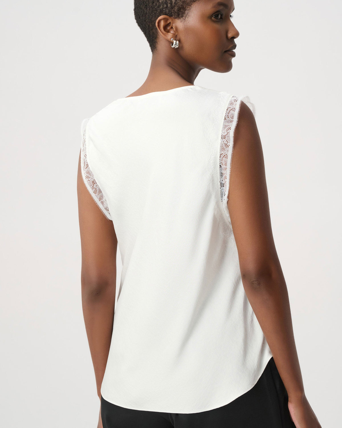 A woman shown from the back wearing a white sleeveless top with delicate lace trim along the shoulder edges, paired with black pants, against a plain light gray studio background.