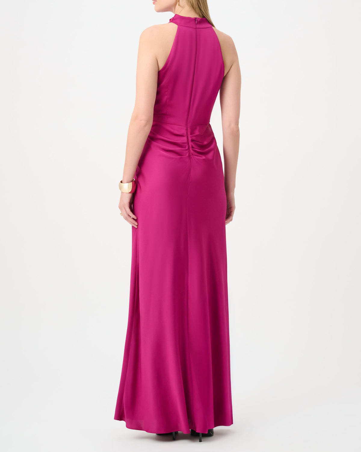 A woman modeling a sleeveless magenta maxi dress from the back, showing a high neckline, back cutout details, and gathered waist, set against a plain white studio background.