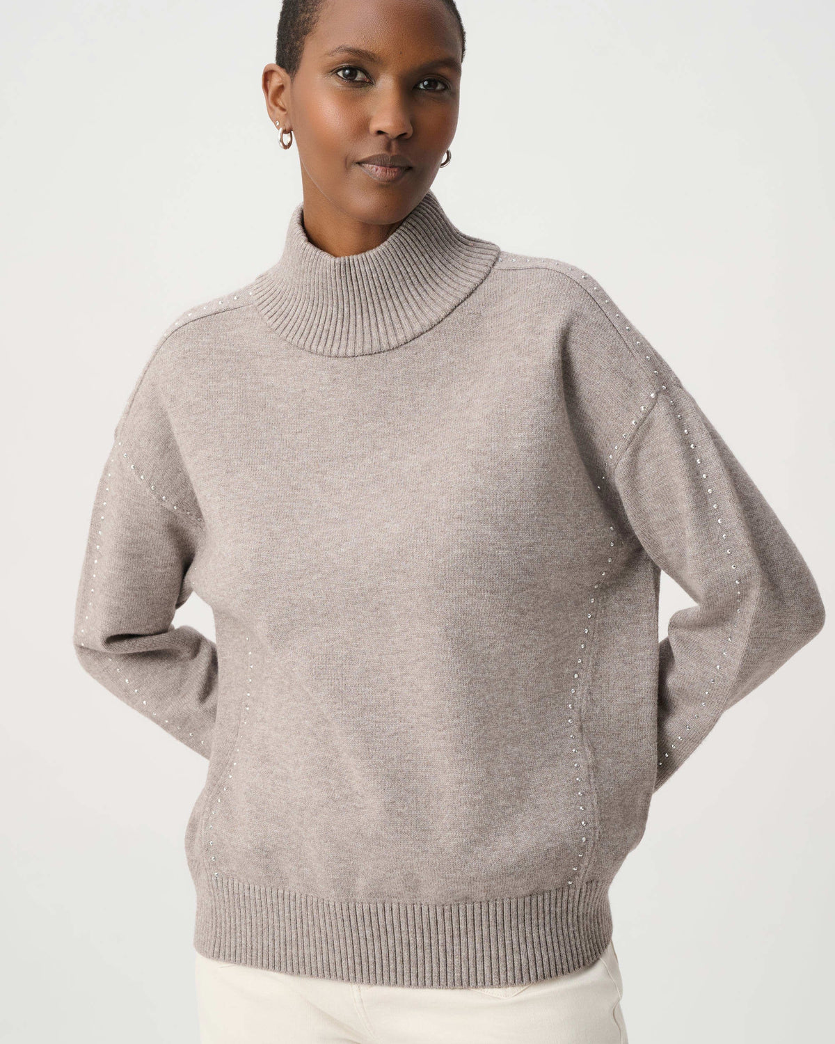 A woman wearing a light taupe knit mock neck sweater with subtle textured details on the sleeves, paired with white pants, against a plain light gray studio background.