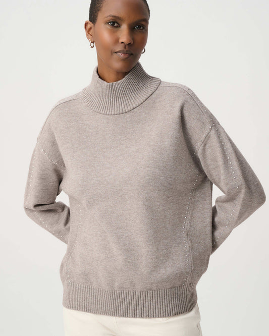 A woman wearing a light taupe knit mock neck sweater with subtle textured details on the sleeves, paired with white pants, against a plain light gray studio background.