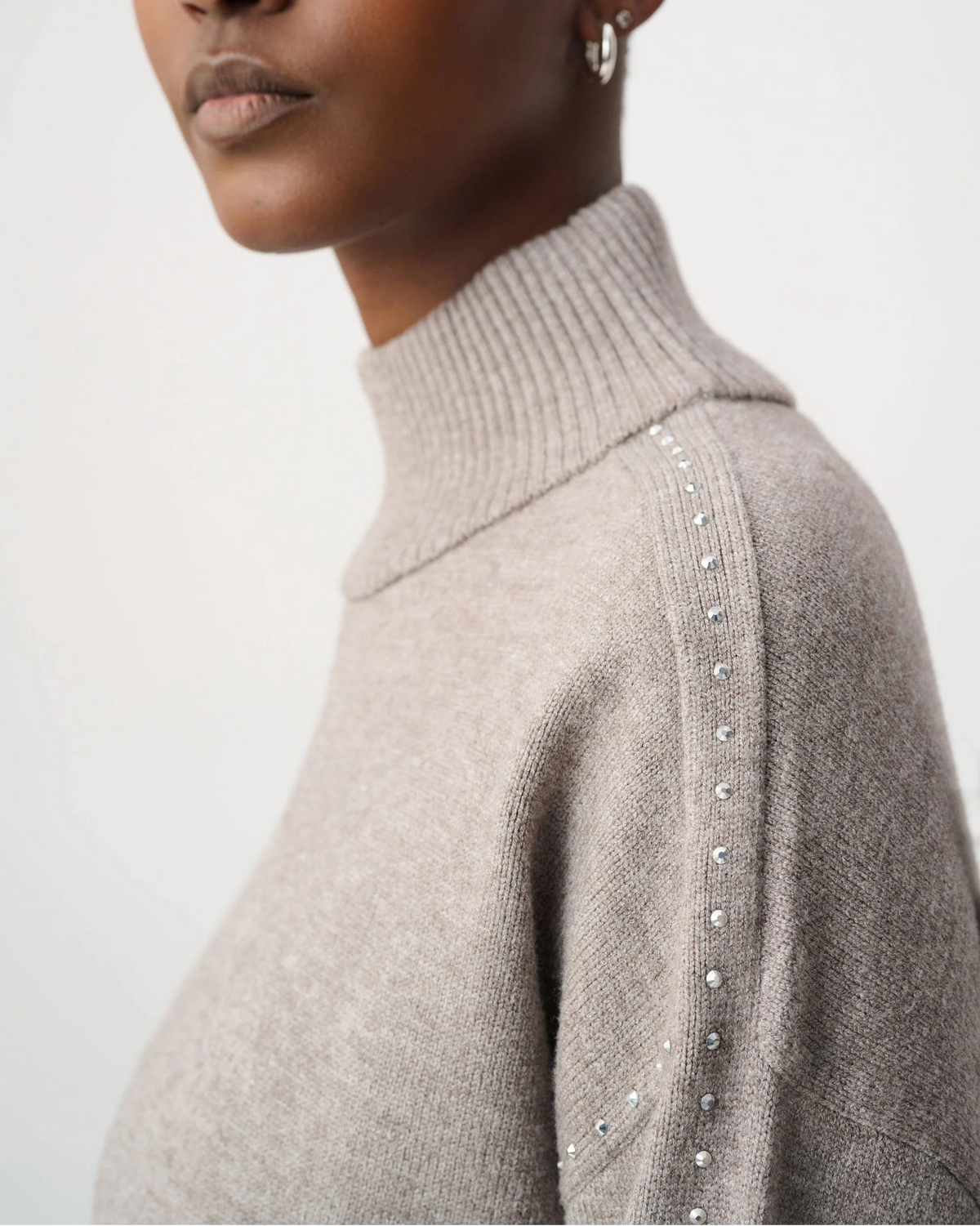 Close-up of a model wearing a light taupe knit turtleneck sweater with silver rhinestone embellishments along the shoulder seam, set against a plain light gray background.