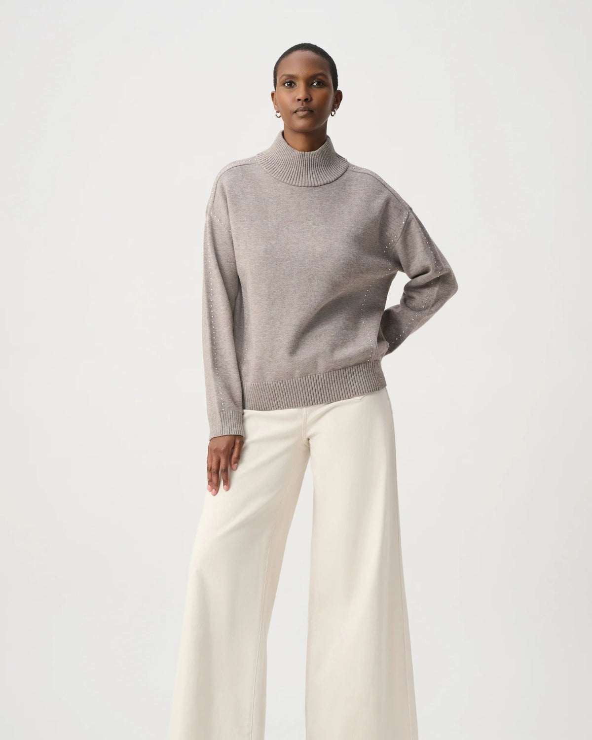 A model stands against a light gray studio background wearing a taupe-gray knit mock-neck sweater and off-white wide-leg trousers.