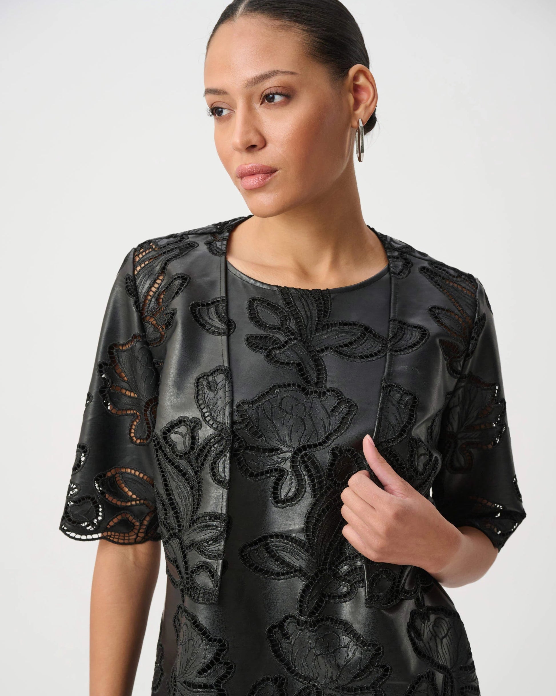 A woman wearing a black short-sleeved dress with intricate floral embroidery and sequined detailing, photographed against a light gray studio background.