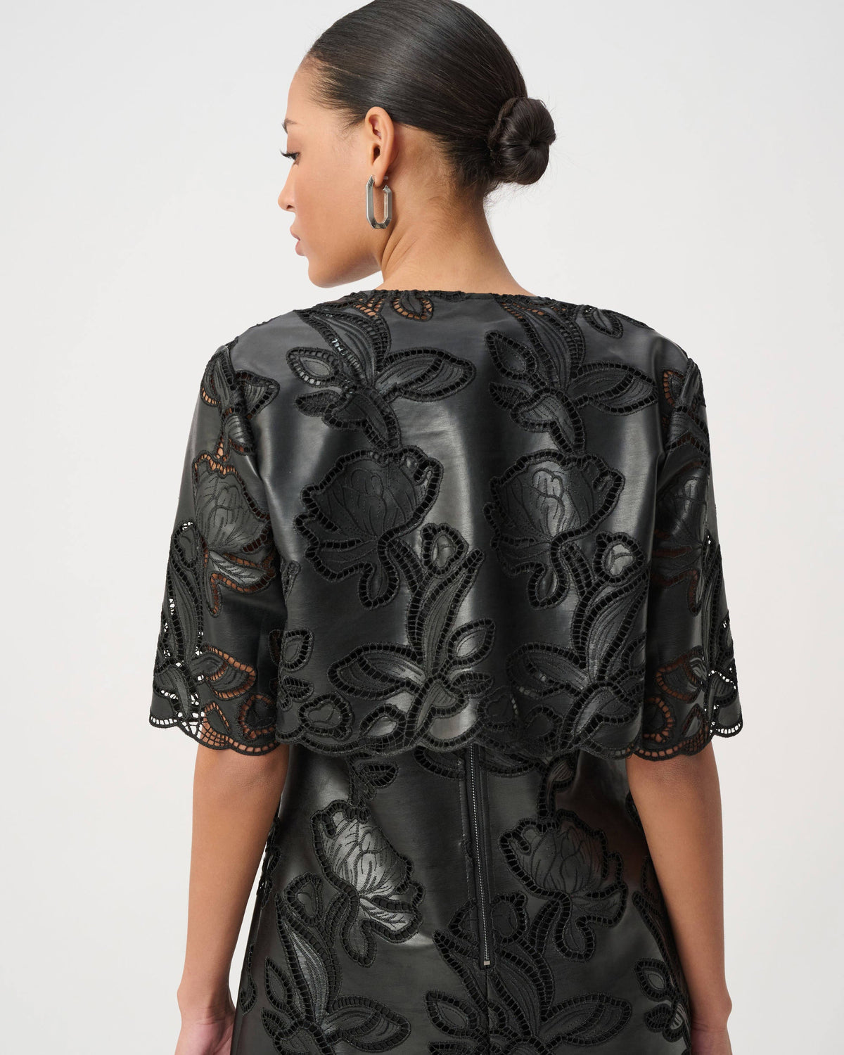 A woman shown from the back wearing a black dress with raised floral embroidery and sheer embroidered sleeves, photographed against a light gray studio background.