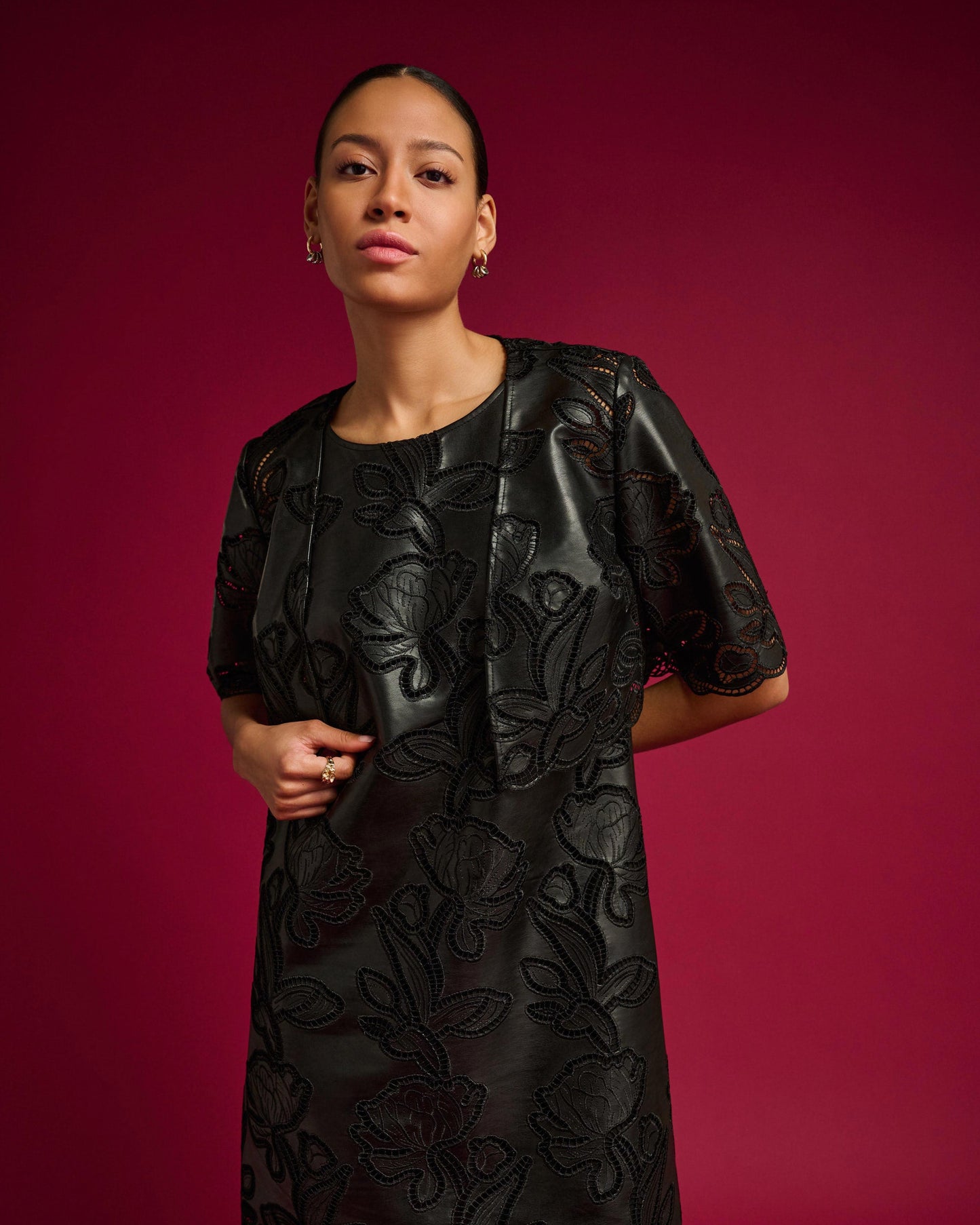 A woman modeling a black short-sleeve dress with a textured, embossed floral pattern against a solid deep magenta background.