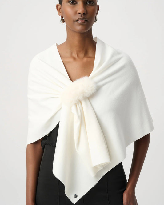 A model wearing a cream-colored wrap shawl with a fluffy pom-pom detail at the center, styled over a black top against a light gray studio background.