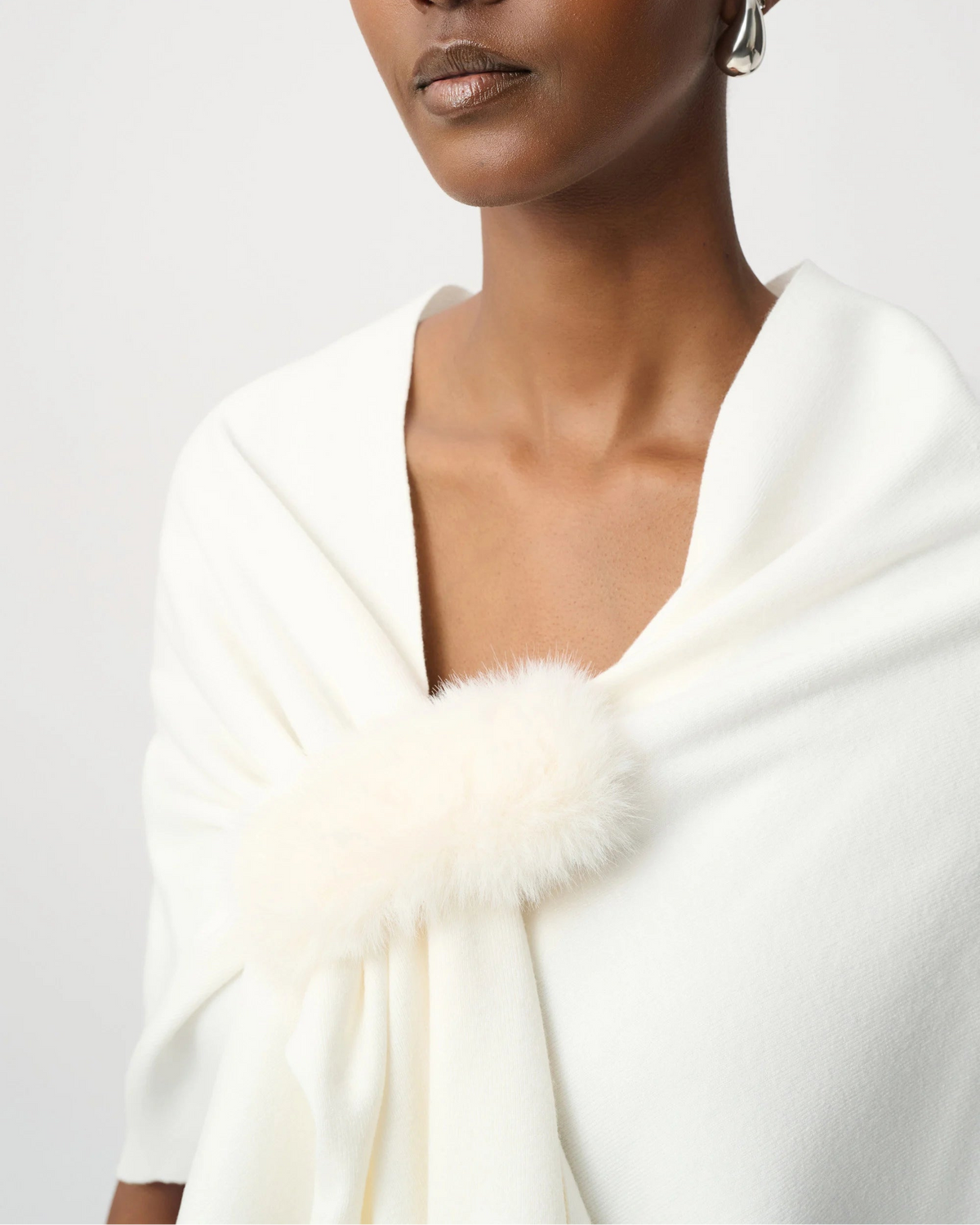 A close-up of a model wearing a white shawl with a fluffy faux-fur trim detail at the neckline, set against a plain light gray background.