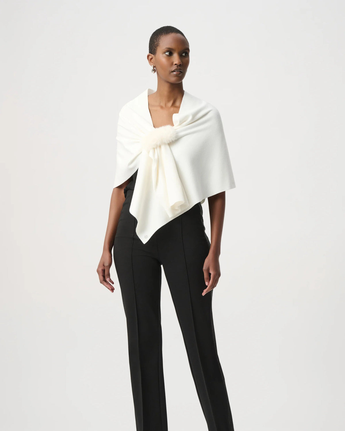 A model wearing a white draped wrap with a large bow at the chest over a black top and black trousers, standing against a light gray studio background.