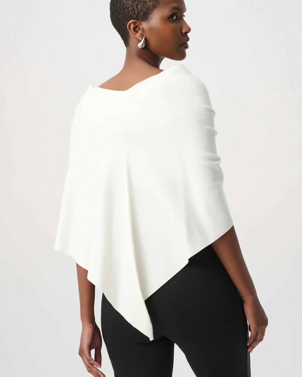 A woman shown from the back wearing a white draped poncho-style top over black pants, photographed against a clean light gray studio background.