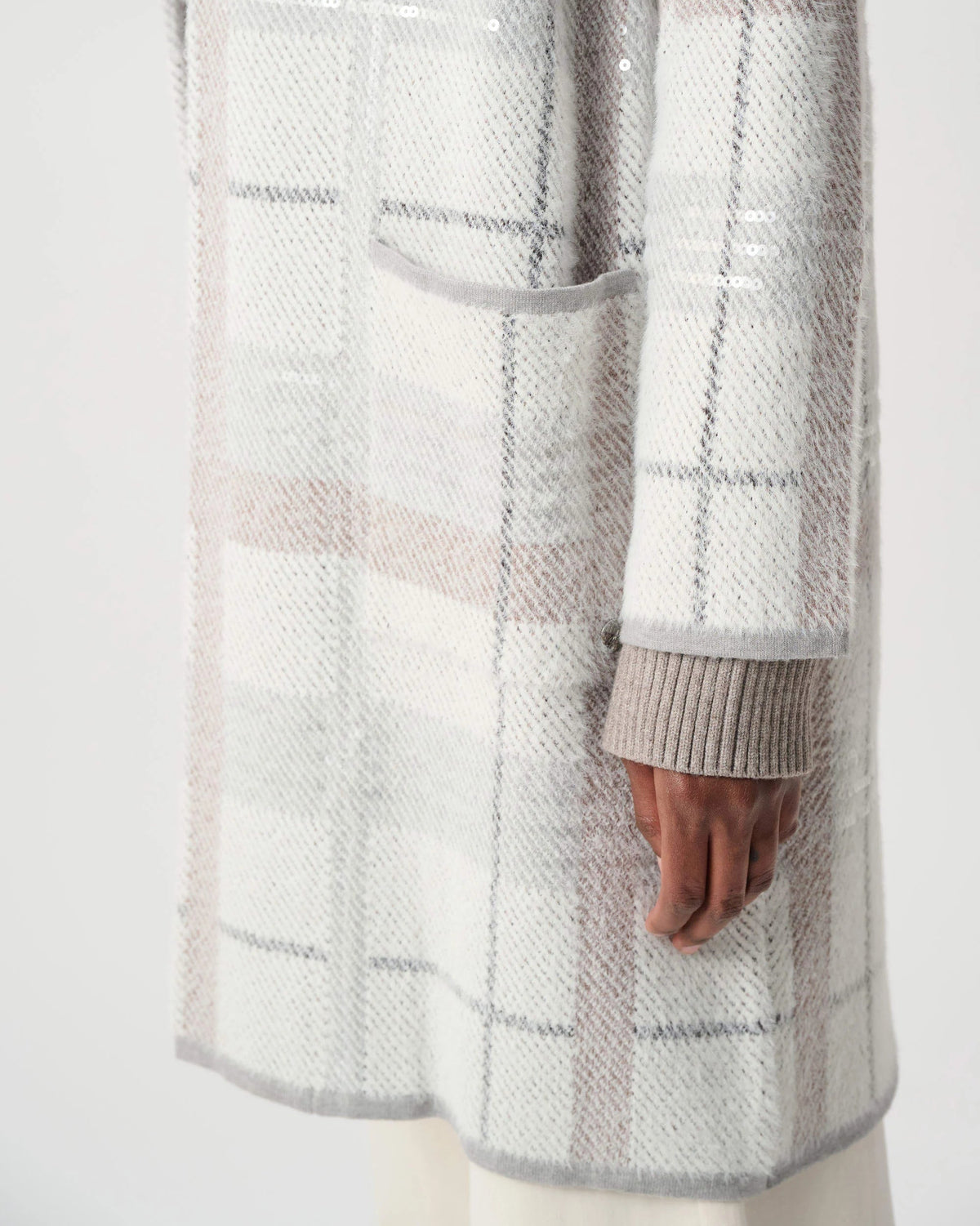 A close-up of a person wearing a textured knit cardigan with a soft plaid pattern in off-white, light gray, and pale pink. The cardigan features a ribbed cuff and a front patch pocket, shown against a plain light background.