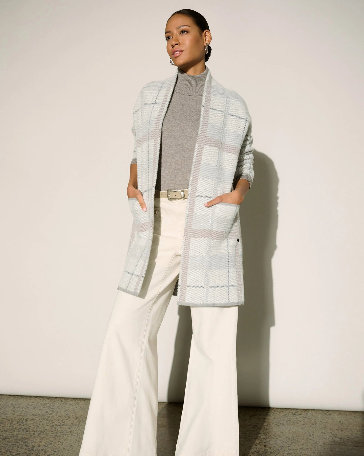A model stands against a plain off-white wall wearing a light gray and white plaid open-front cardigan over a gray mock-neck top and cream wide-leg pants.