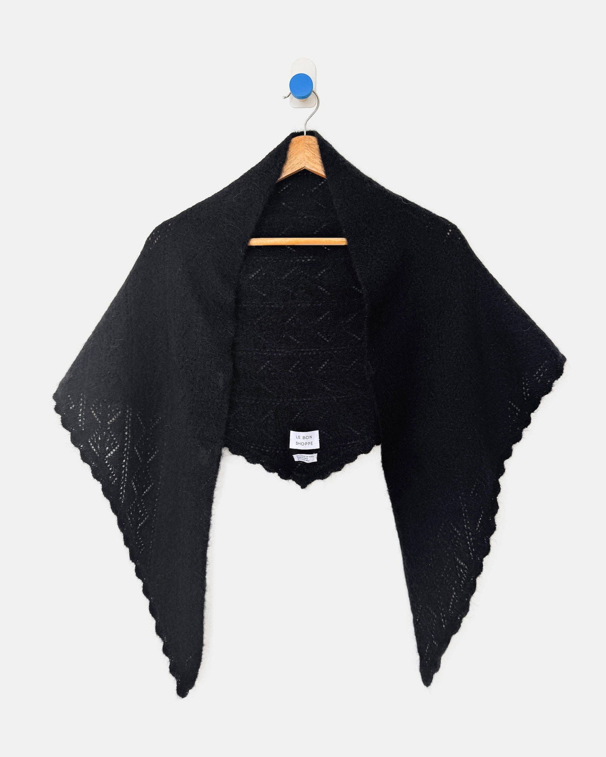 A black knit shawl with scalloped edges hanging from a wooden hanger against a light gray background.