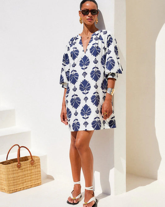 Woman wearing a white dress with blue leaf pattern standing against a white wall.