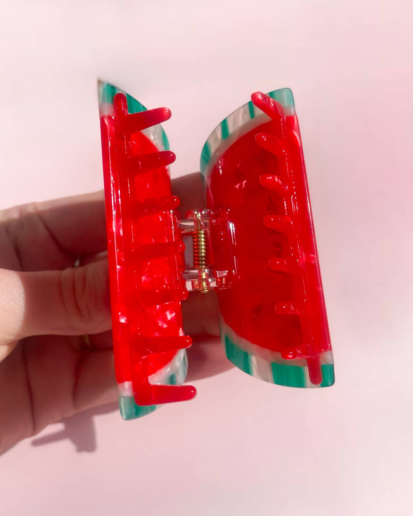 Watermelon-shaped hair clip with green and red striped design held by a hand on a pink background