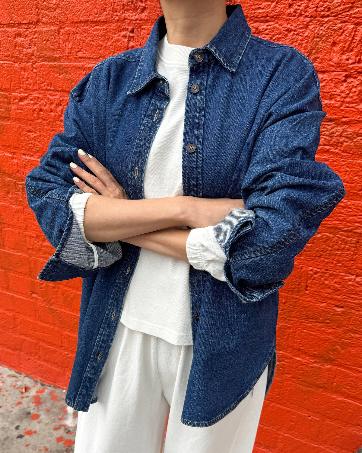 A person wearing an oversized dark blue denim button-up shirt over a white t-shirt and white pants, standing with arms crossed in front of a bright orange-red brick wall.
