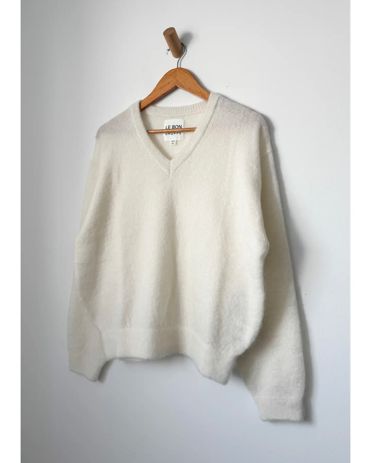 Le Bon Shoppe James V-Neck Sweater in snow hanging on a wooden hanger against a white wall.