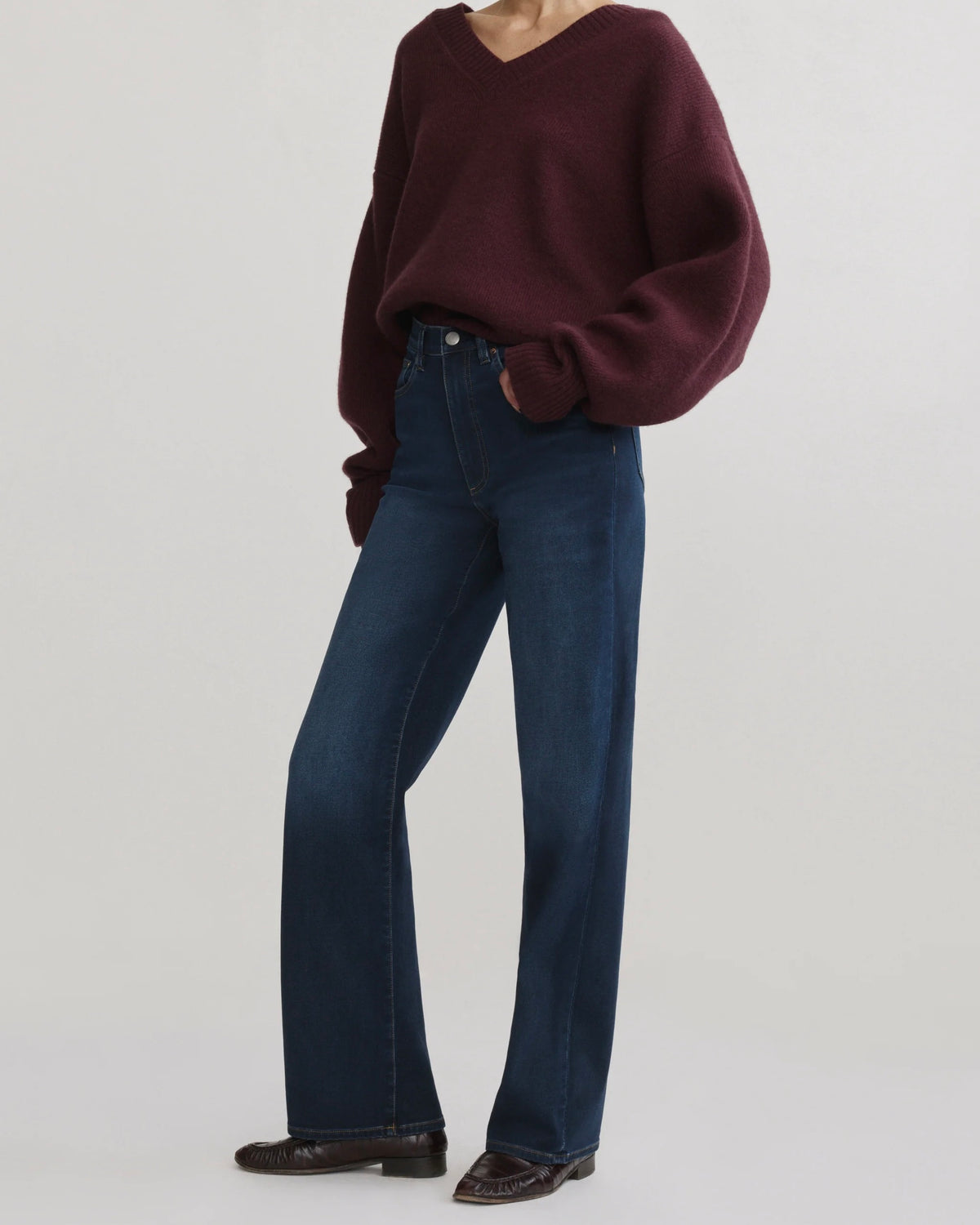 A model wearing a dark burgundy V-neck knit sweater and dark blue wide-leg jeans, paired with dark brown leather shoes, against a plain light gray studio background.