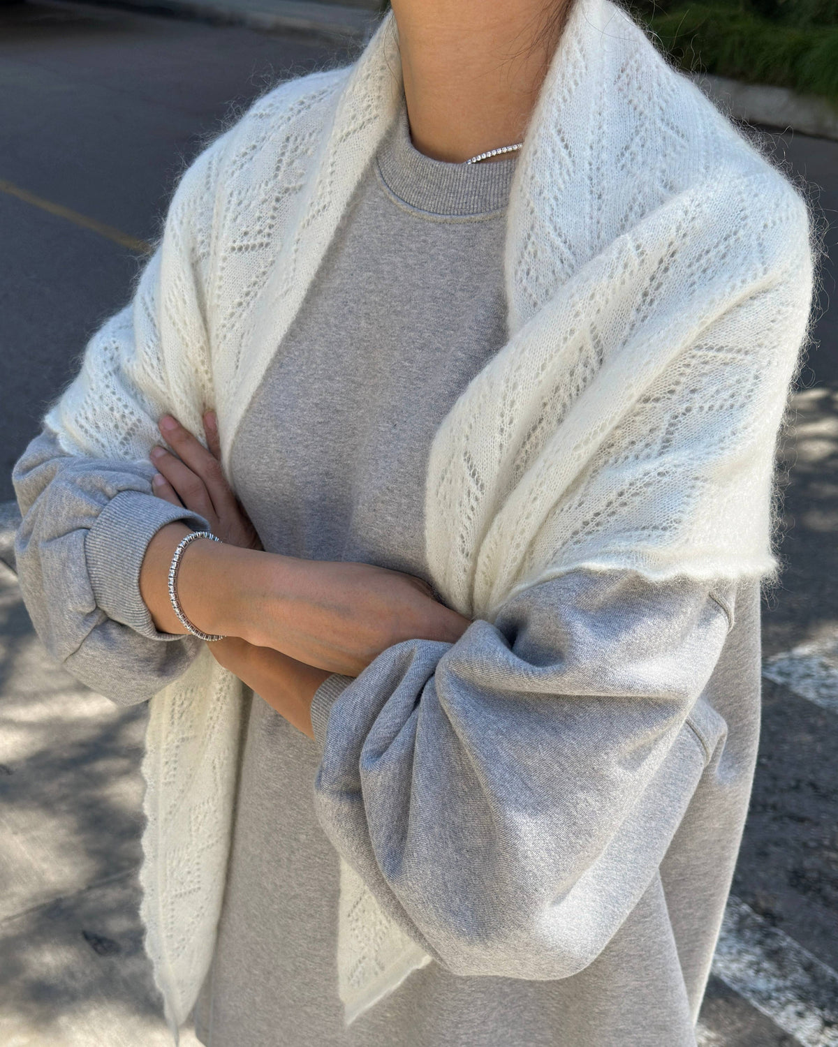 A person outdoors wearing a light gray crewneck sweatshirt with a cream-colored cable-knit shawl draped over the shoulders.