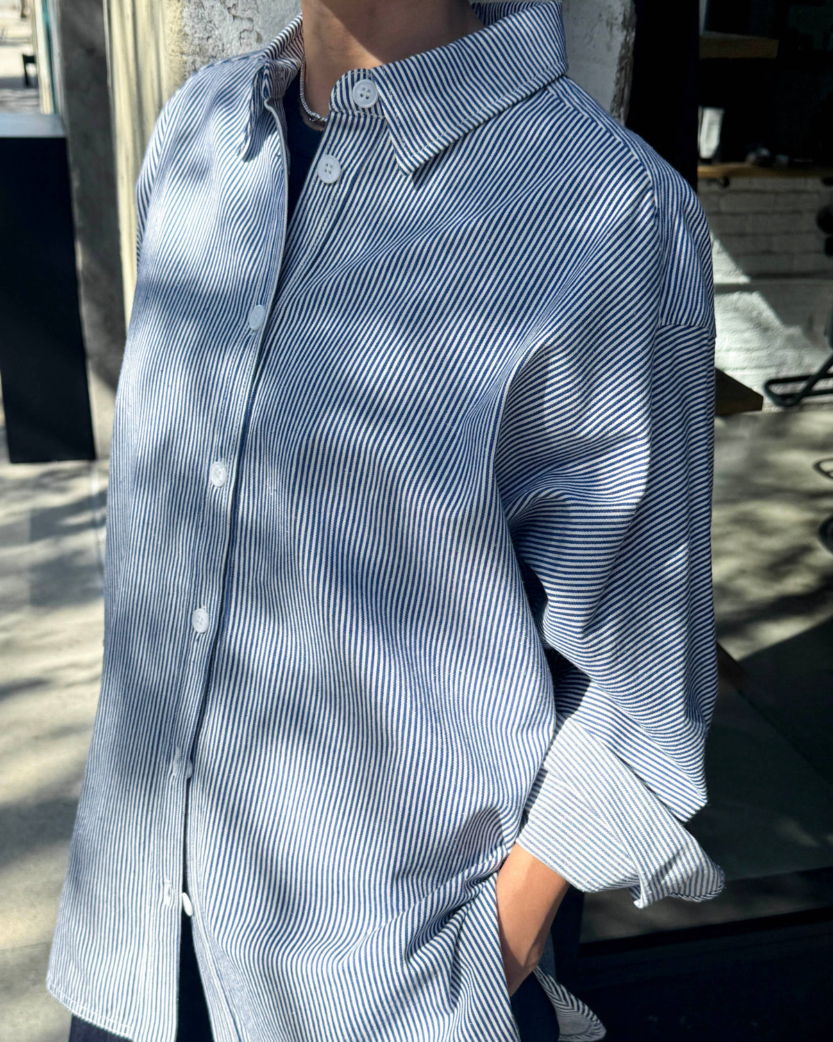 A person wearing a light blue and white vertically striped button-down shirt with dark trousers, standing outdoors in dappled sunlight.