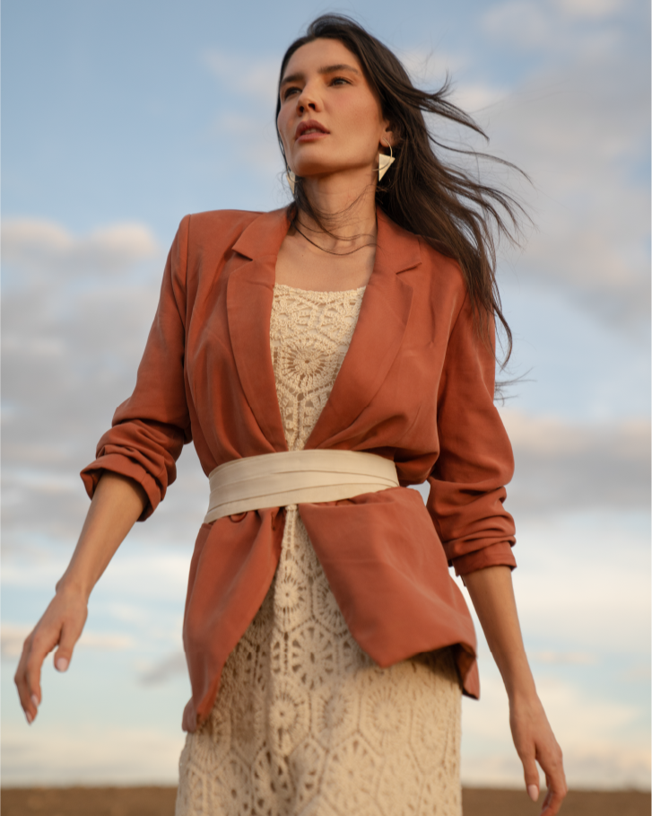 Woman wearing a rust-colored jacket over a lace dress against a cloudy sky.