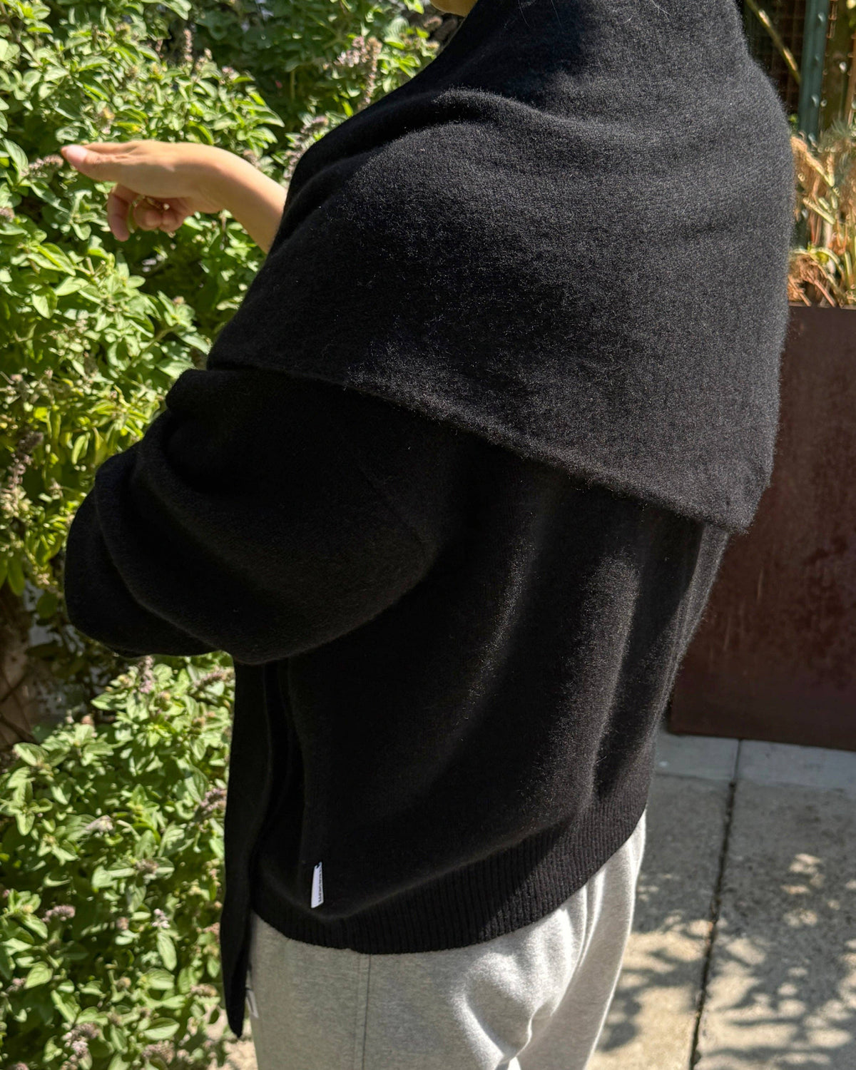 A person outdoors wearing a black knit shawl draped over their shoulders and a black cardigan, paired with light gray sweatpants. The background features green foliage and a concrete walkway.