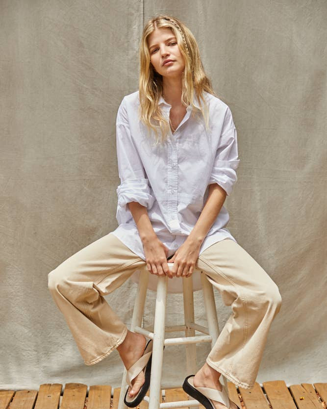 Woman sitting on a stool wearing a white shirt and beige pants against a plain background