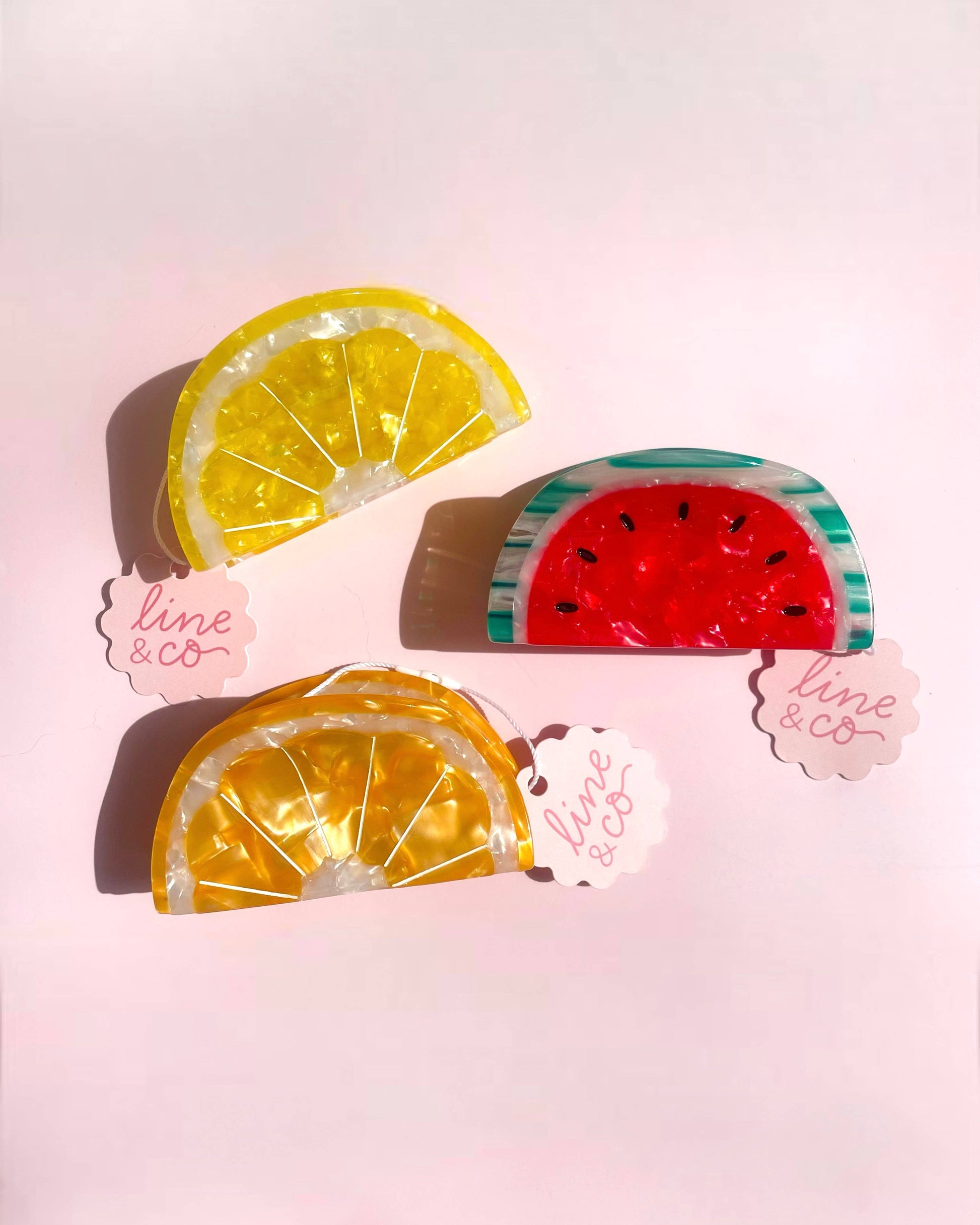Three hair clips shaped like lemon, watermelon, and orange slices on a pink background with 'line & co' branding.