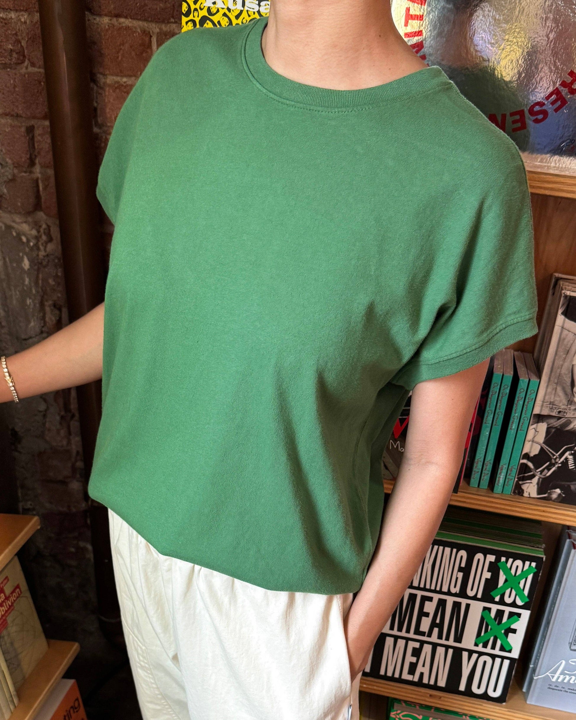 Person wearing a green oversized t-shirt and white pants in front of a bookshelf.
