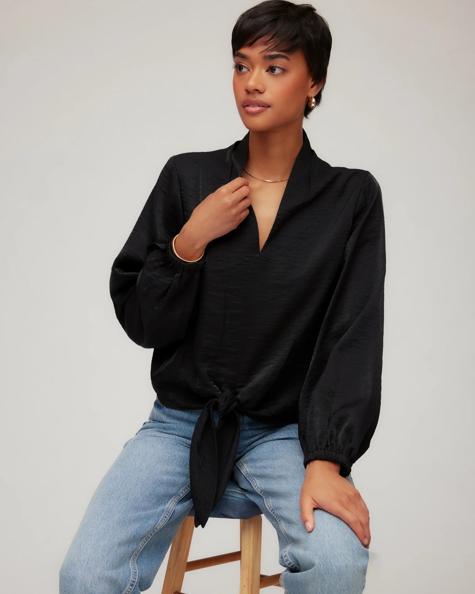 Woman wearing a black blouse and blue jeans sitting on a stool against a plain background