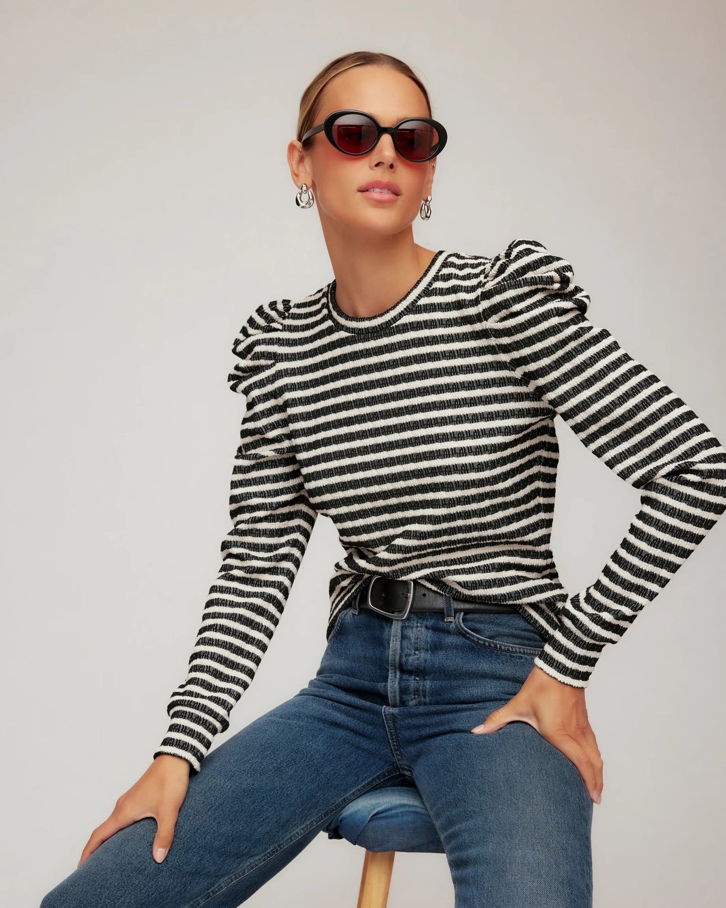 Woman wearing a black and white striped sweater and blue jeans sitting on a stool against a plain background.