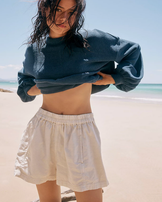 Model Wearing Free People Get Free Poplin Pull Nilla Cream Shorts Wearing A Blue Sweater Showing Her Stomach On The Beach