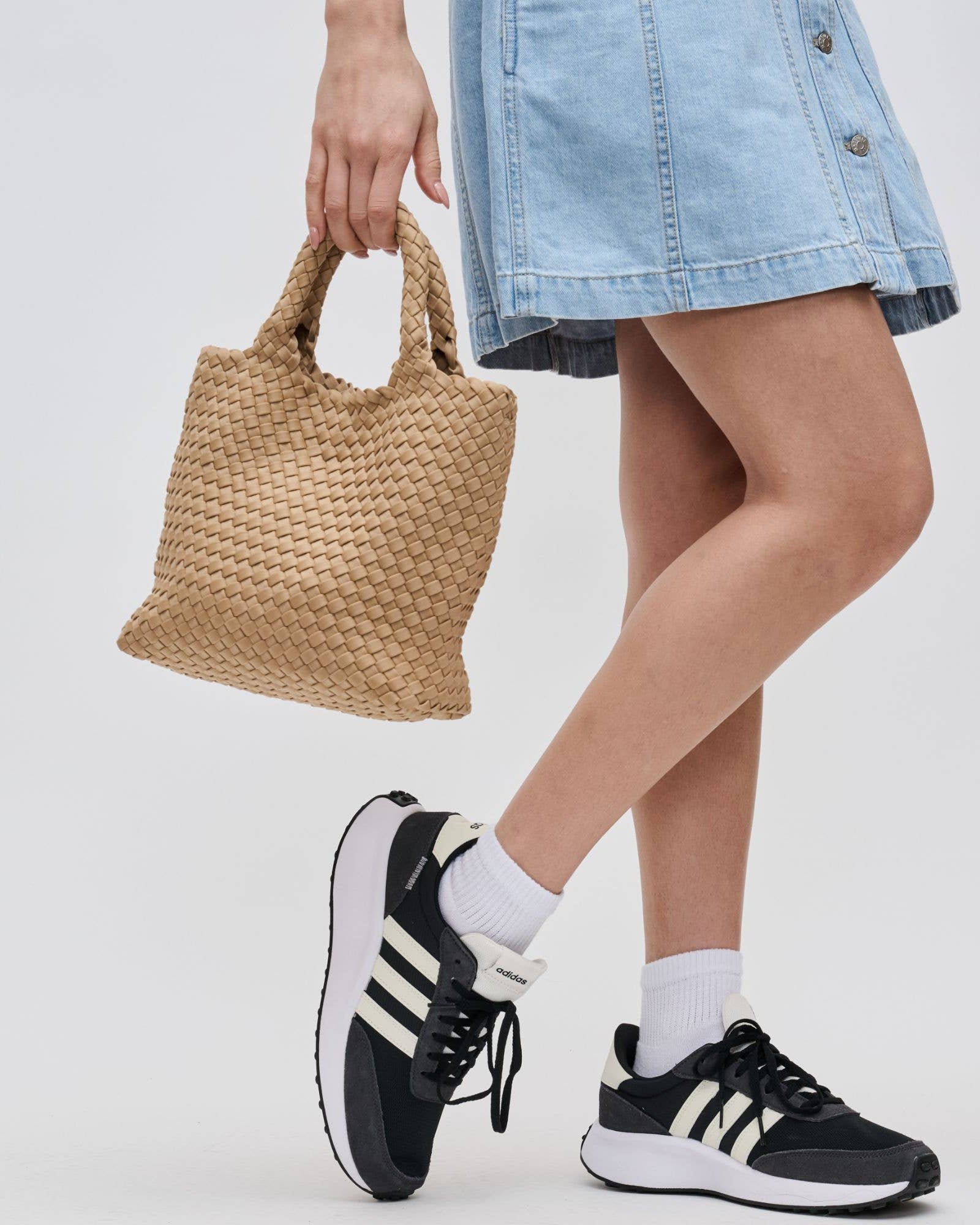 Person wearing black Adidas sneakers with white stripes, holding a woven bag, and wearing a denim skirt.