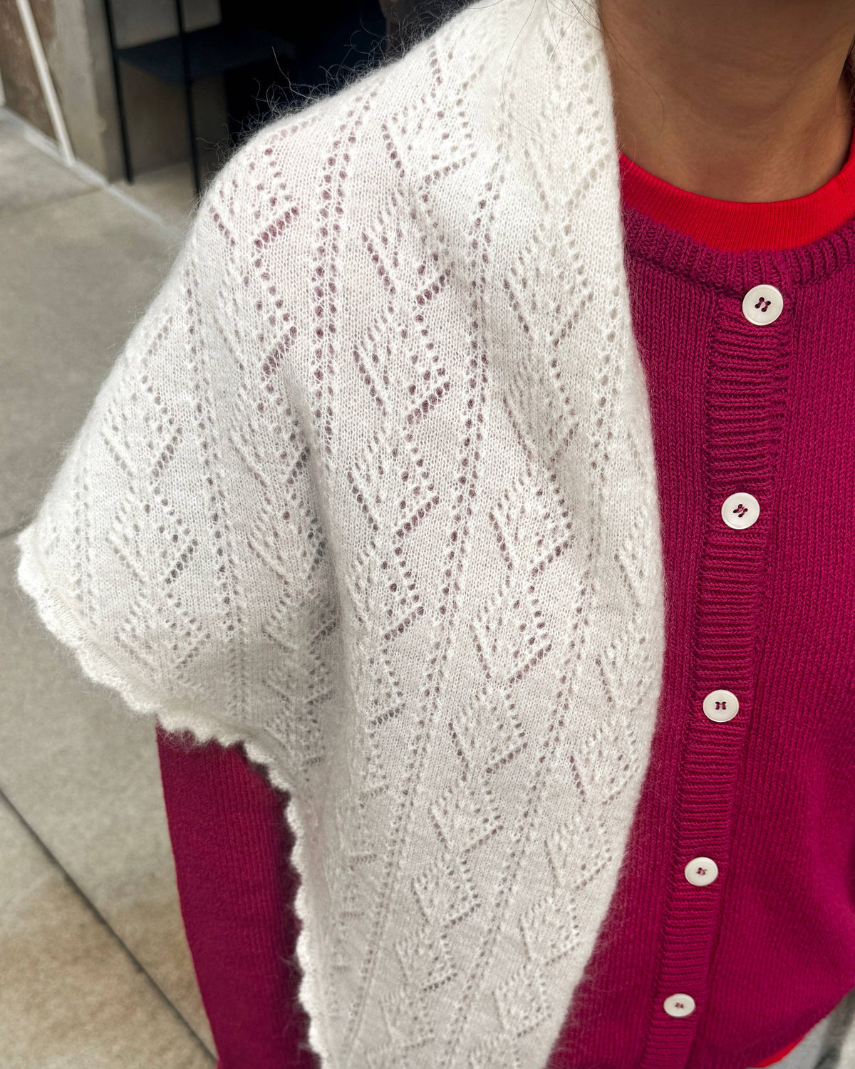 A person wearing a white open-knit shawl or capelet over a bright pink button-front cardigan and gray pants, photographed indoors with a tiled floor visible in the background.