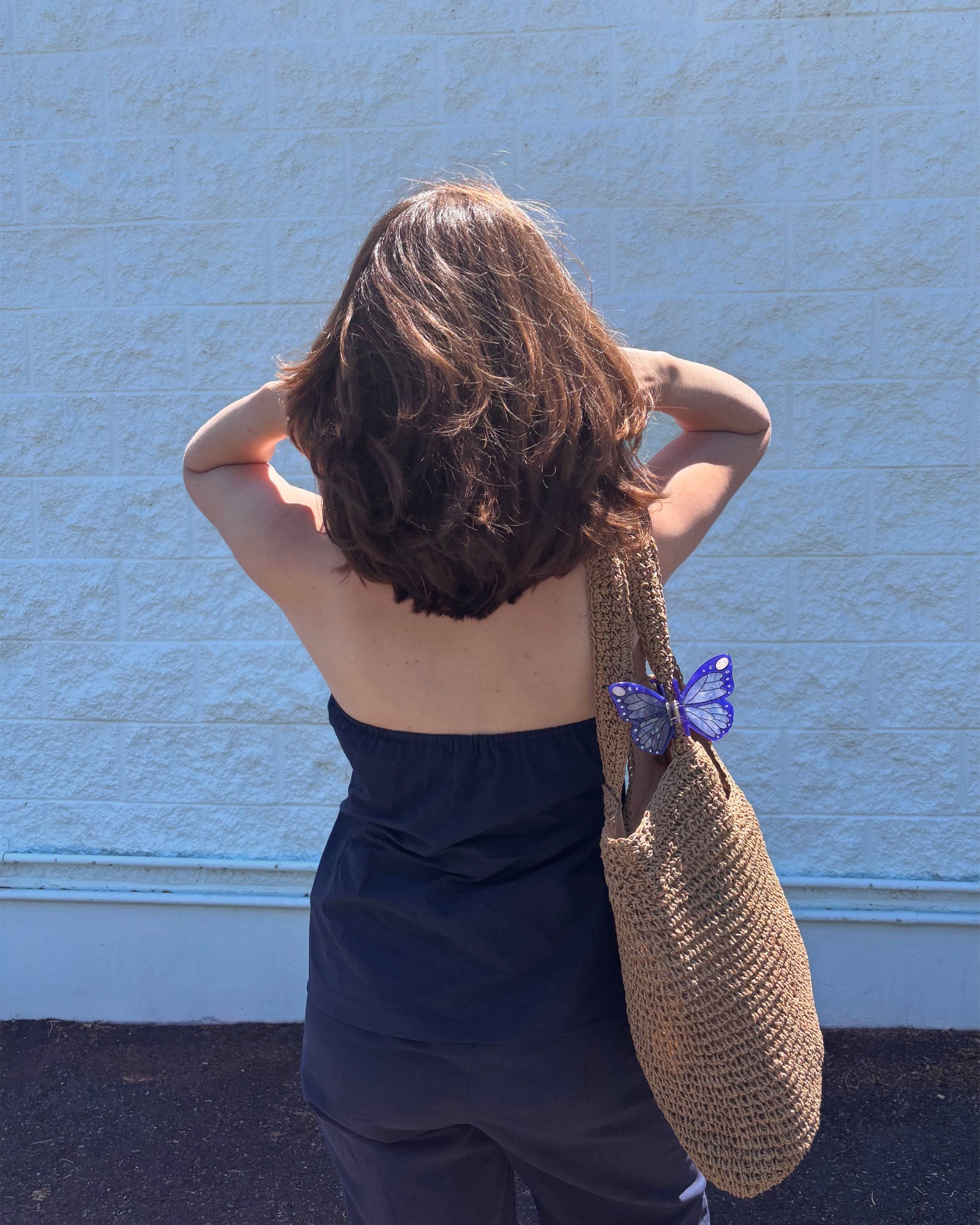 Person with a blue strapless top and dark pants holding a woven bag against a blue wall.
