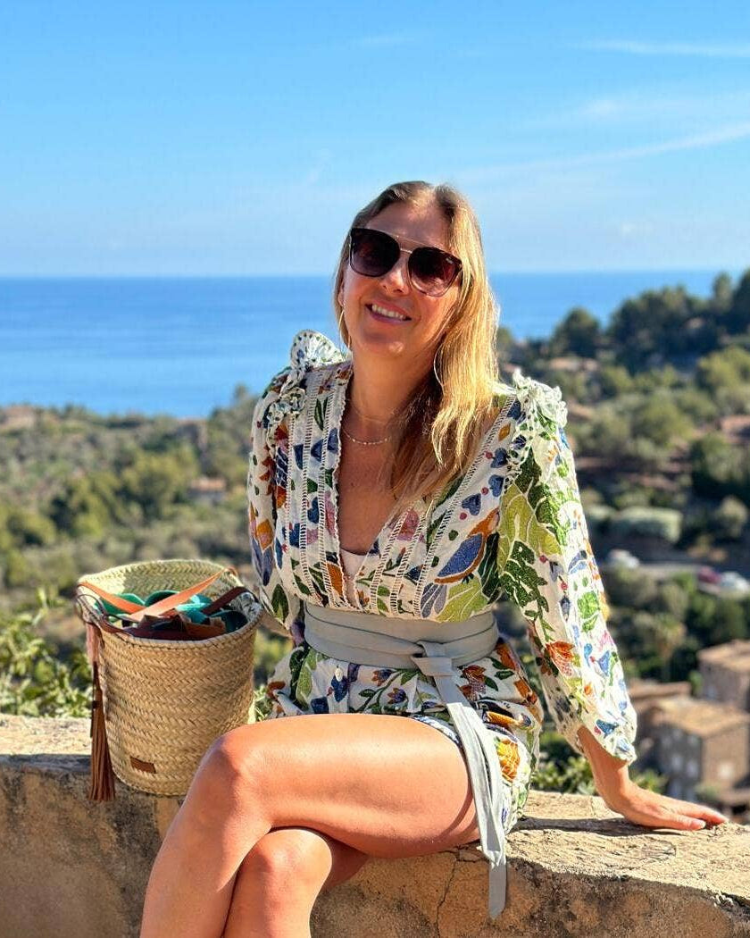 Woman in a floral dress sitting on a ledge with a scenic background of ocean and trees.