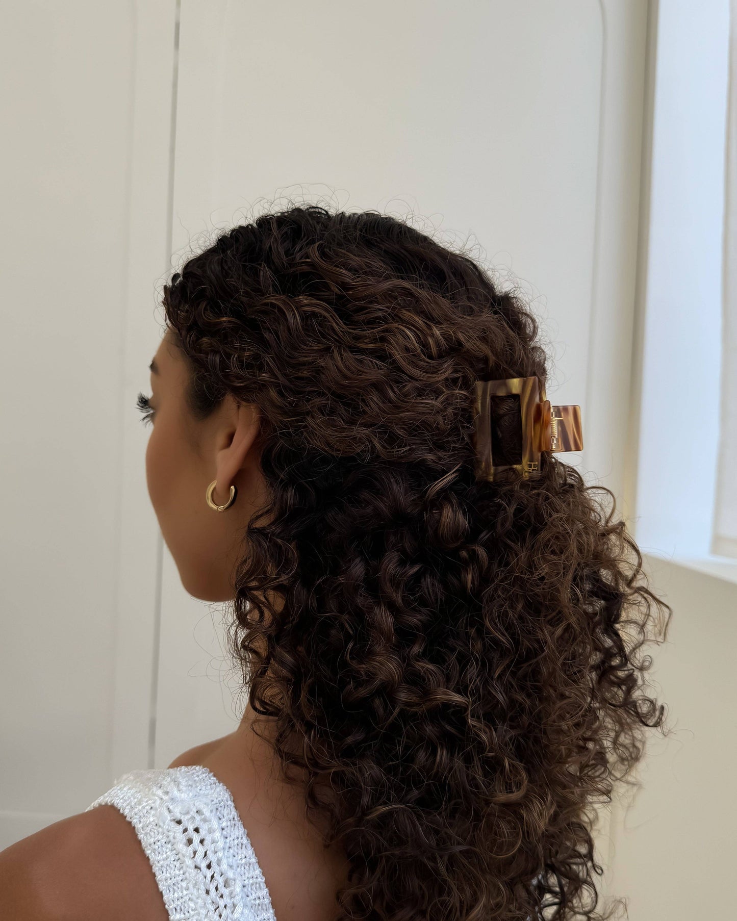 Woman with curly hair styled with the HoneyLux Ecofriendly Acetate Midi Clip in the color Hazel Honey wearing a white sleeveless top, in a softly lit room