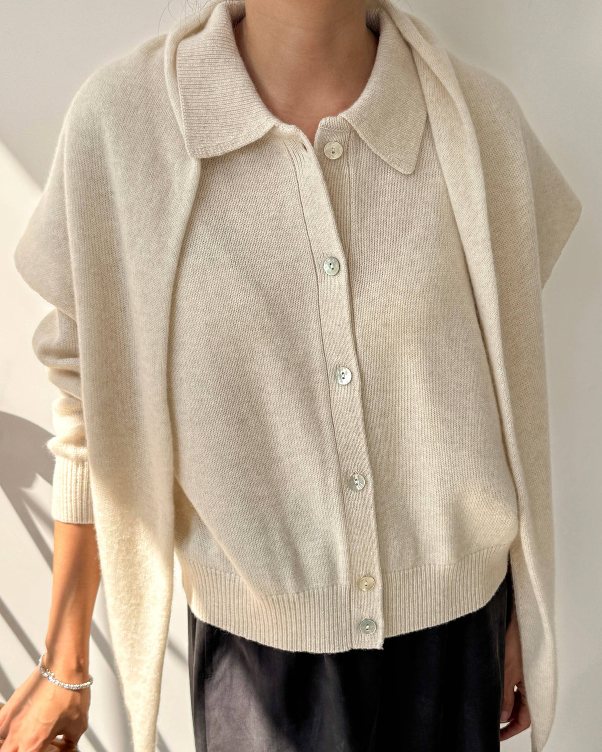A person wearing a cream-colored knit polo cardigan with wide draped sleeves and dark charcoal pants, standing indoors against a light wall.