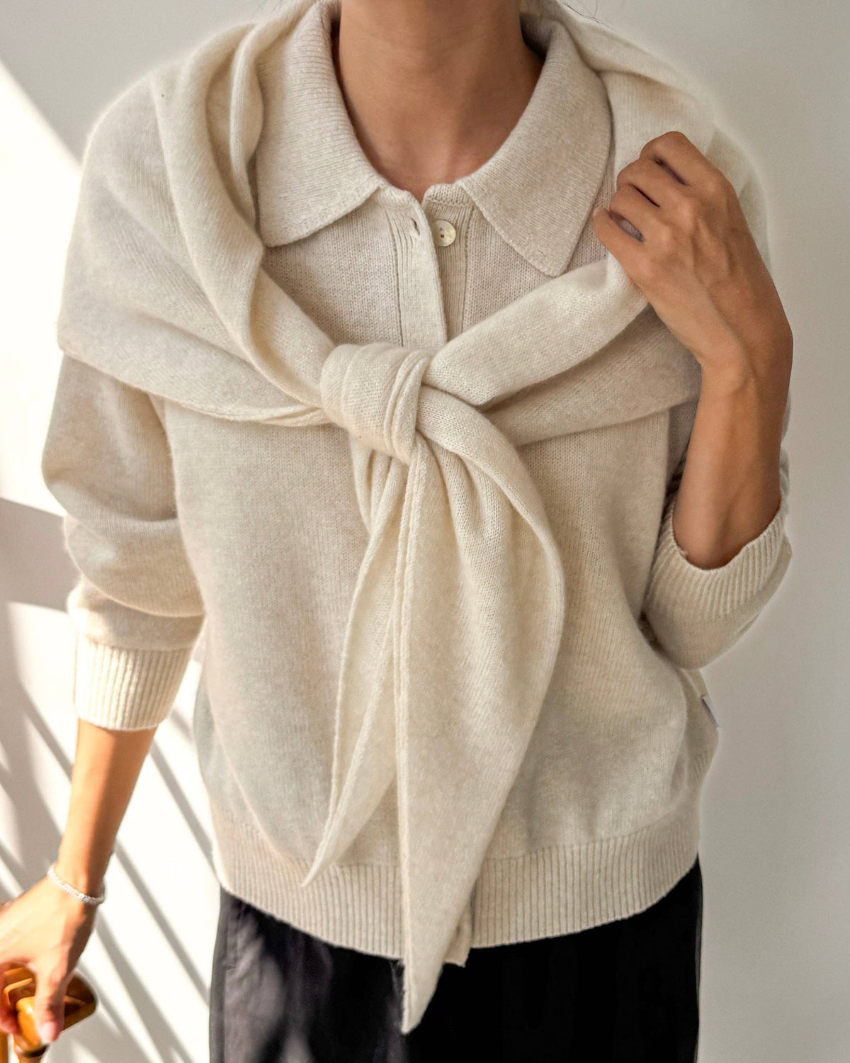 A person wearing a cream-colored knit cardigan sweater with a matching knotted scarf, paired with black pants, in a brightly lit indoor setting.