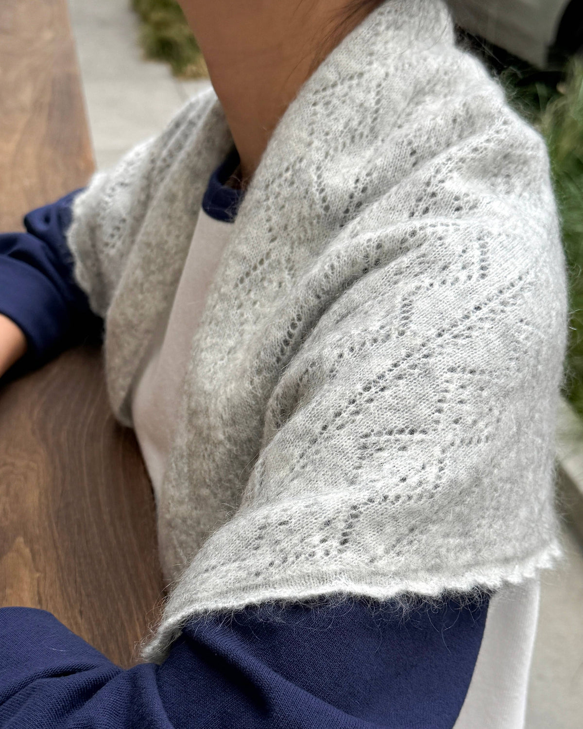 A close-up of a person wearing a light gray open-knit shawl or cardigan over a navy and white long-sleeve shirt, seated at a wooden table.