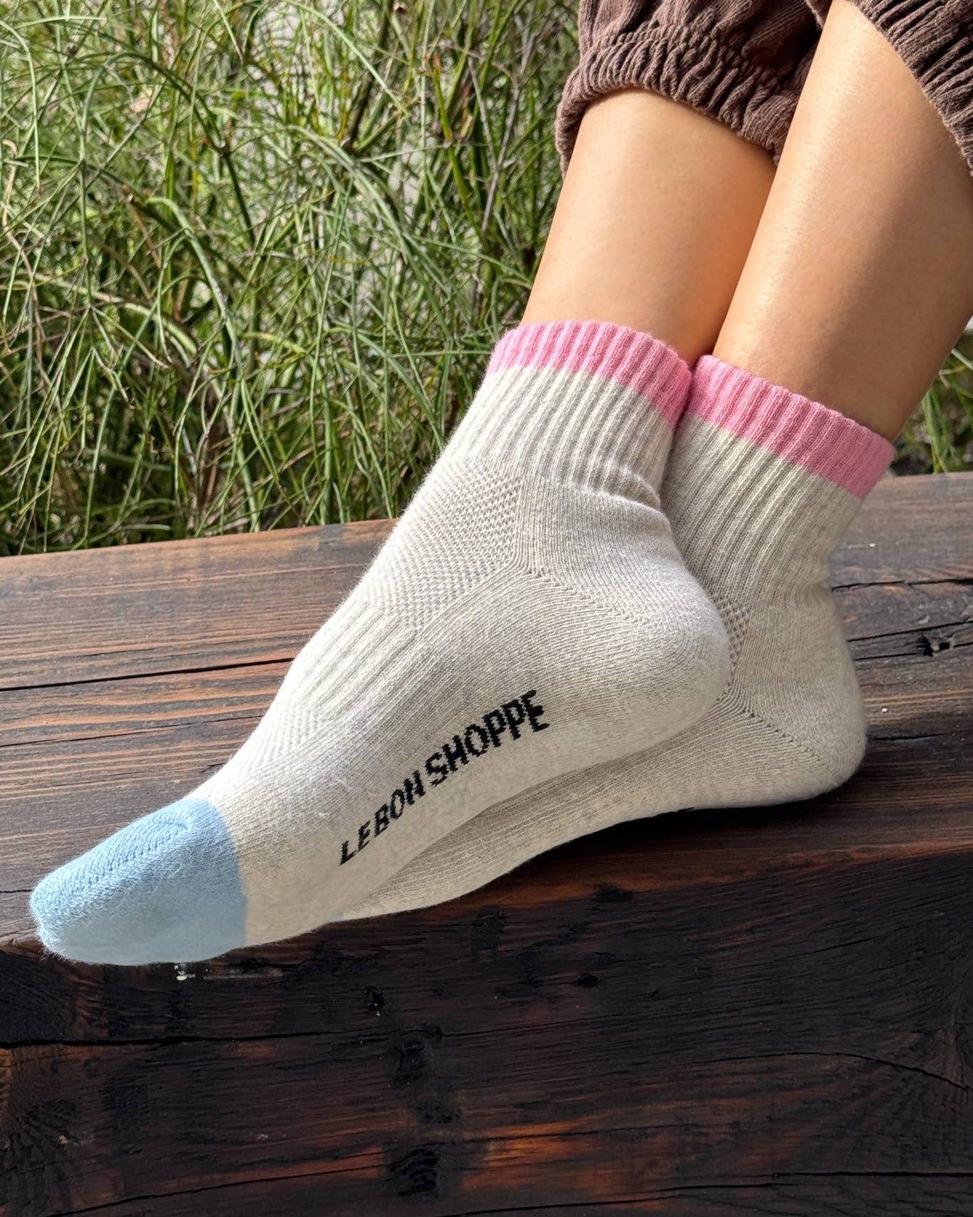White socks with pink and blue toes worn by a person sitting on a wooden bench with grass in the background.