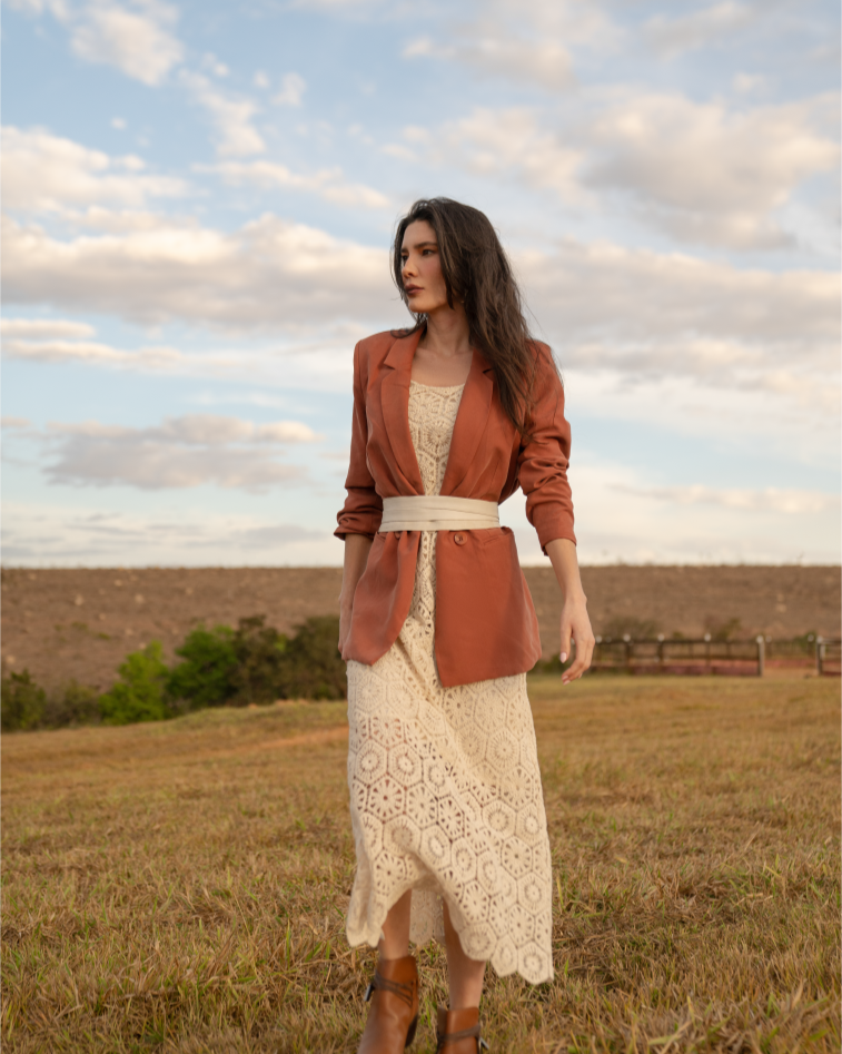 Woman in a rust-colored jacket and white dress standing in a field with a blue sky.