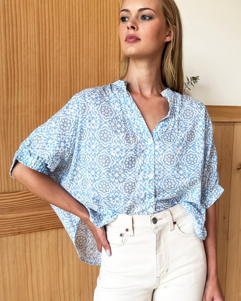 Model wearing Emerson Fry Mandarin Collar Top in Blue Geo Flower color wearing white pants standing in front of a brown wall